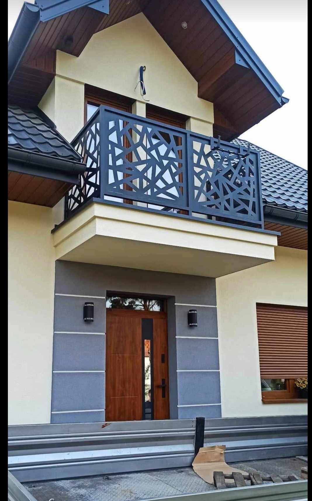 Nowoczesna, ażurowa balustrada balkonowa w geometryczne wzory, zamontowana na jasnej elewacji domu z drewnianą stolarką okienną i drzwiami wejściowymi.