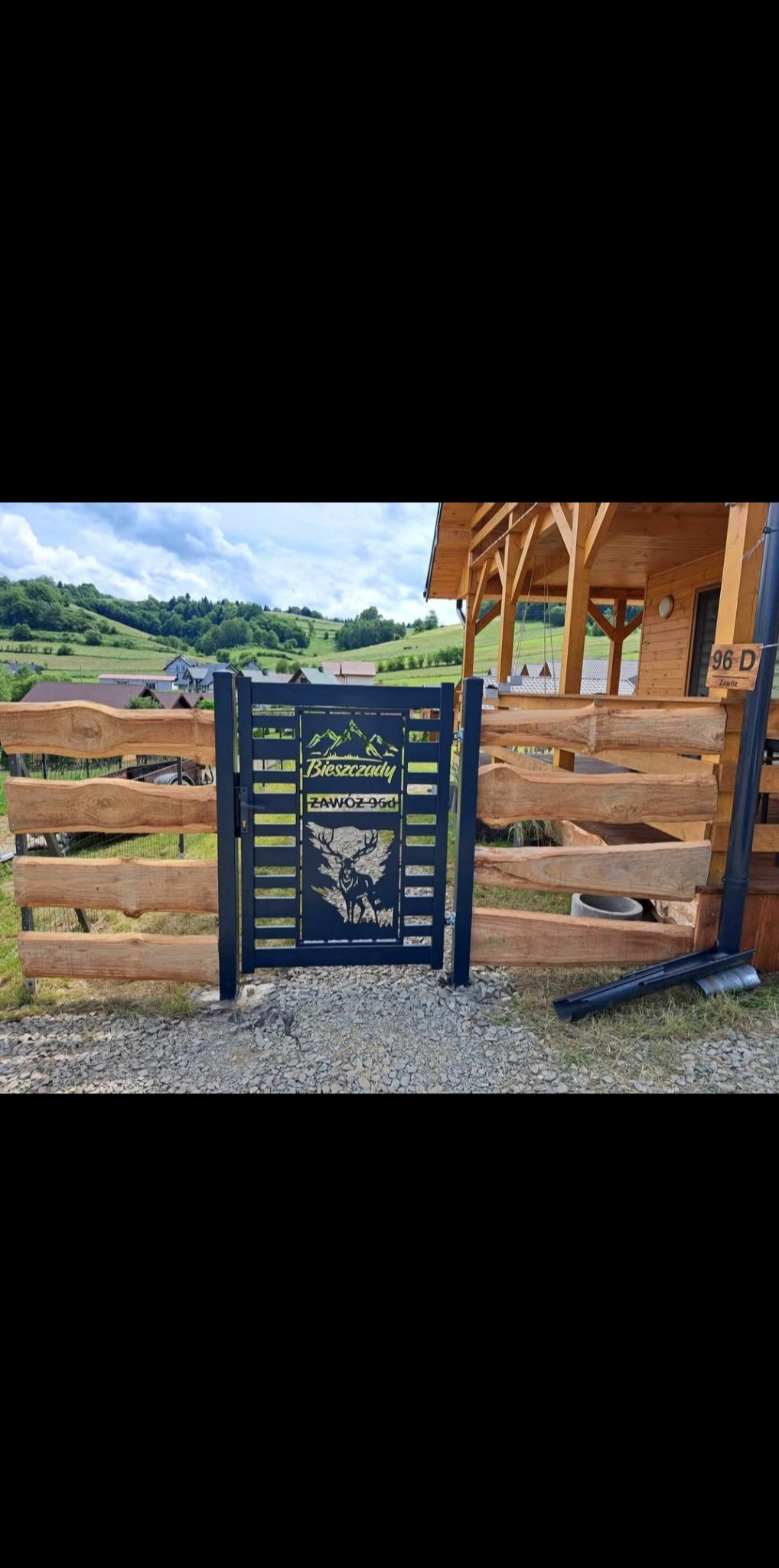 Czarna, metalowa furtka z motywem gór i jelenia, wkomponowana w drewniane ogrodzenie na tle pagórkowatego krajobrazu. Furtka z napisem 'Bieszczady'.