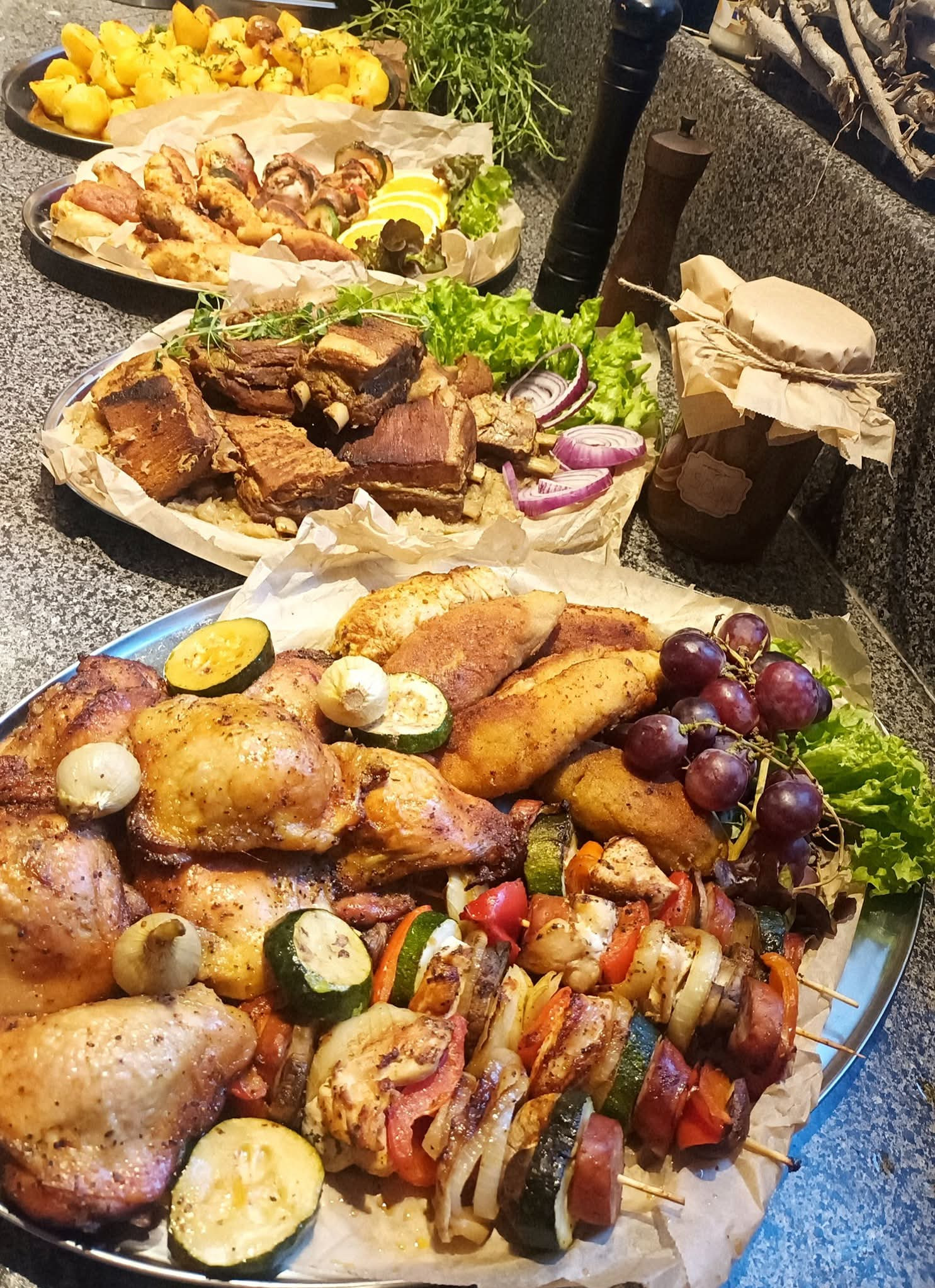 Urozmaicony catering: pieczone mięsa, szaszłyki warzywno-mięsne, ziemniaki, sałata i owoce na srebrnych tacach, w rustykalnej aranżacji.