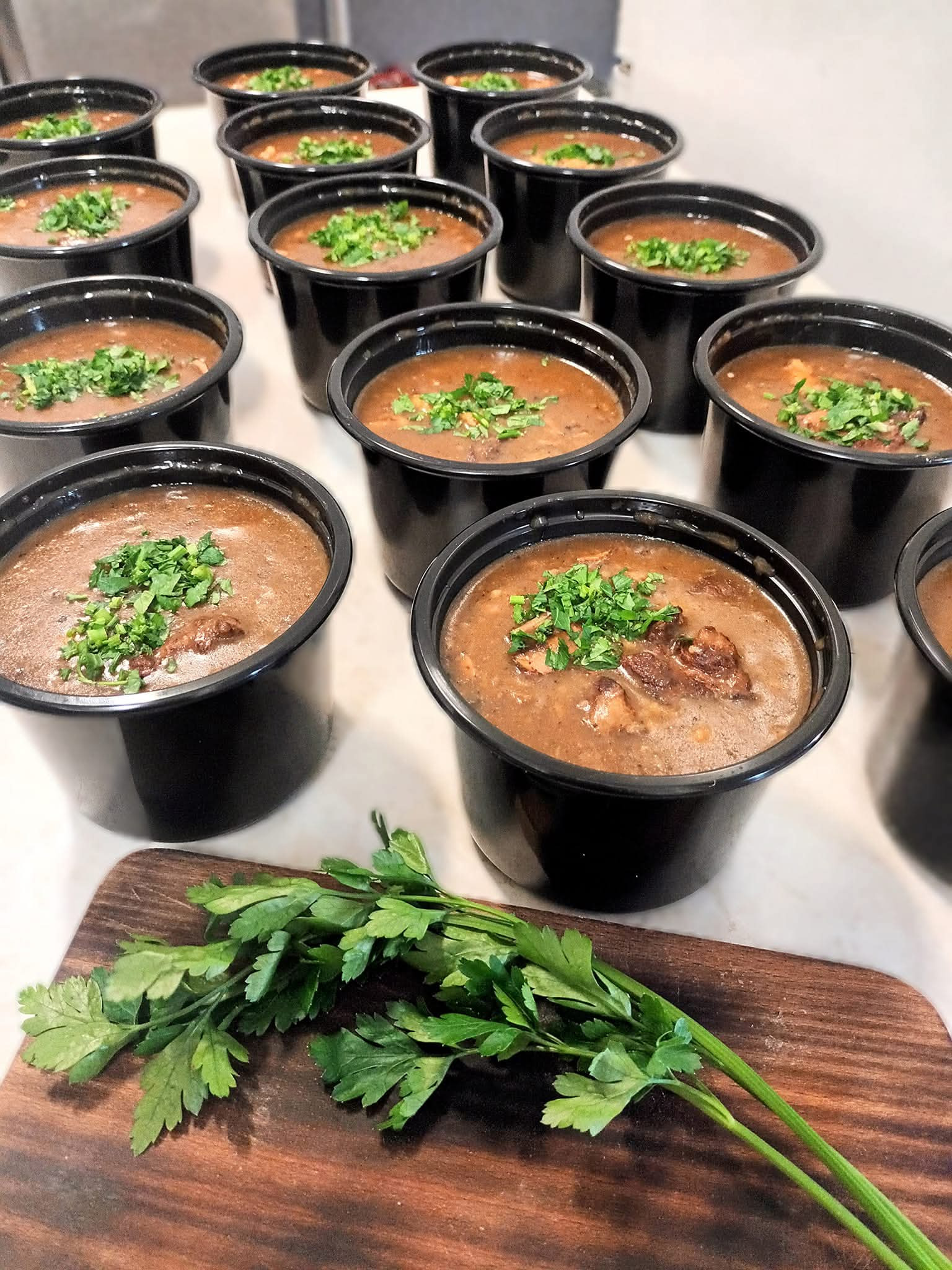 Porcje gulaszu w czarnych pojemnikach, udekorowane świeżą natką pietruszki, ułożone na blacie obok drewnianej deski z zieloną pietruszką. Gotowe dania cateringowe.