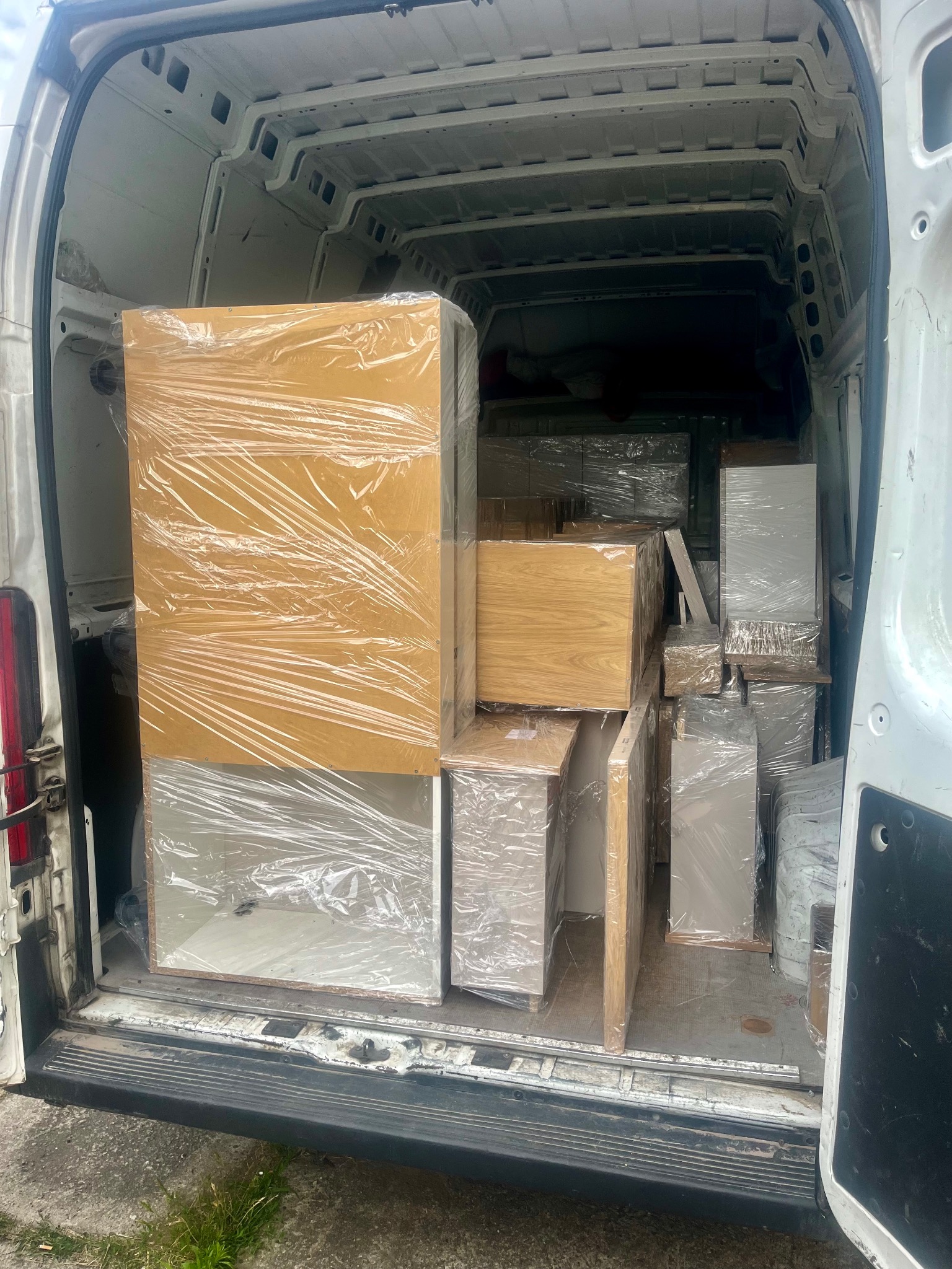 Wnętrze białego busa wypełnione meblami zabezpieczonymi folią stretch, gotowymi do transportu. Widok z tyłu pojazdu, otwarte drzwi.