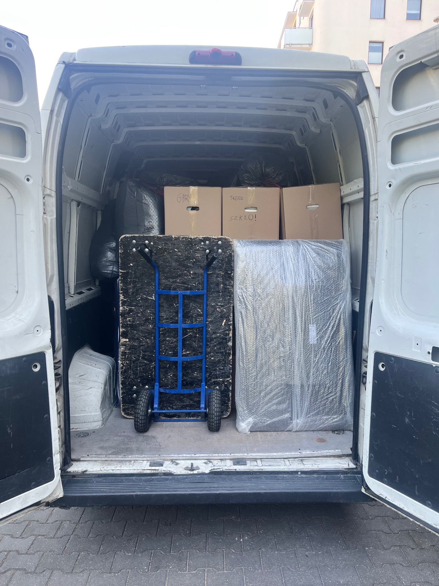Wnętrze białego busa załadowane kartonami, wózkiem transportowym i meblem owiniętym folią stretch, gotowe do przewozu na tle budynku mieszkalnego.