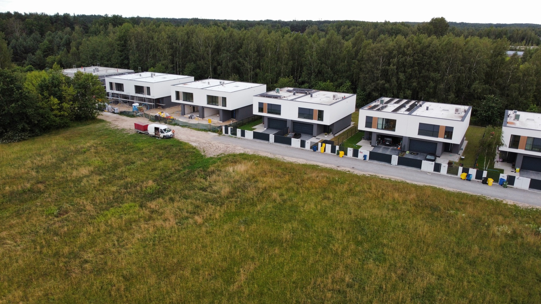 Nowoczesne, dwukondygnacyjne domy z płaskimi dachami, jeden w trakcie budowy, widziane z lotu ptaka na tle lasu i łąki, z zaparkowanym busem budowlanym.