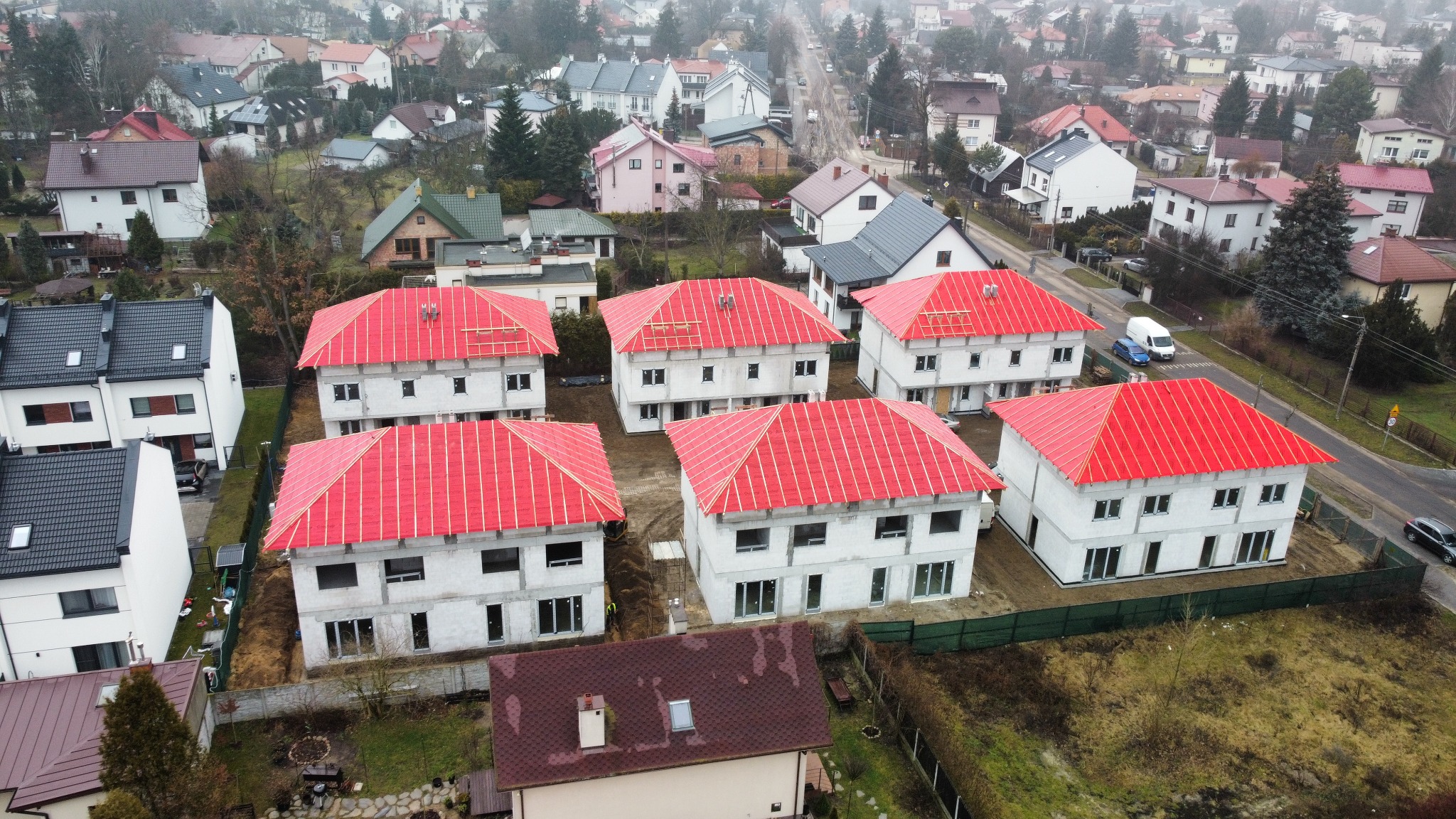 Osiedle domów w trakcie budowy z czerwonym pokryciem dachowym, widok z góry. Domy w stanie surowym zamkniętym, otoczone zielenią i zabudową jednorodzinną.