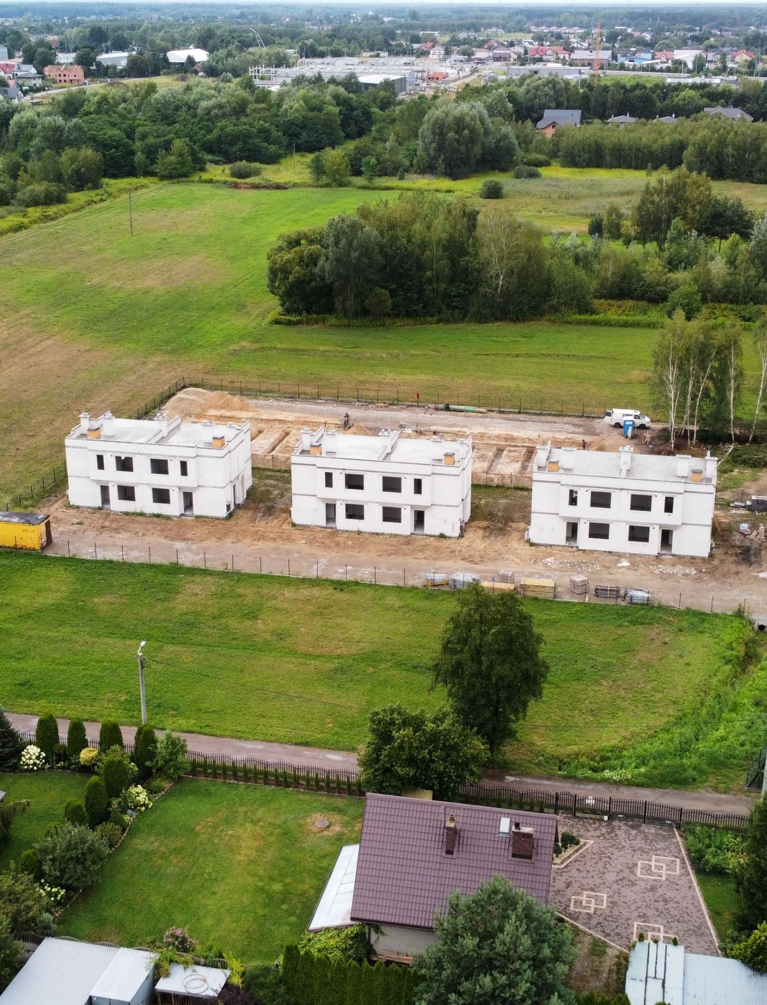 Panorama z lotu ptaka: trzy identyczne, dwukondygnacyjne domy w stanie surowym na zielonym terenie. Widoczne fundamenty i częściowo wymurowane ściany.