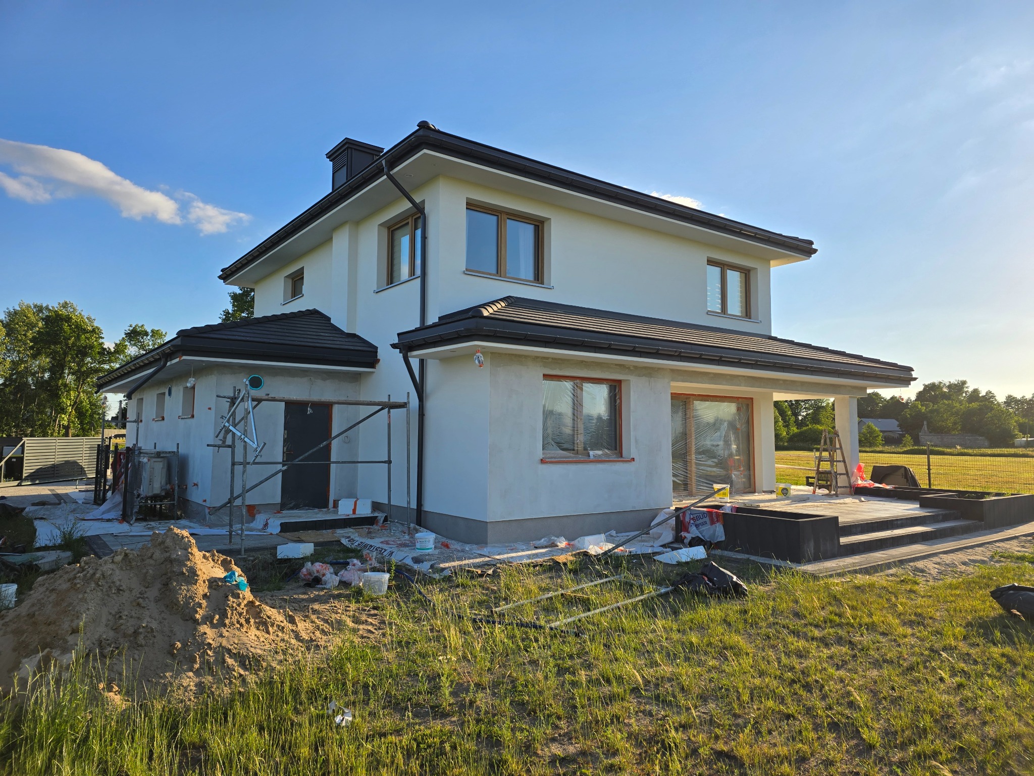 Dwukondygnacyjny dom z białymi ścianami w trakcie wykończenia, widoczne rusztowania i teren przygotowany pod ogród, błękitne niebo w tle.