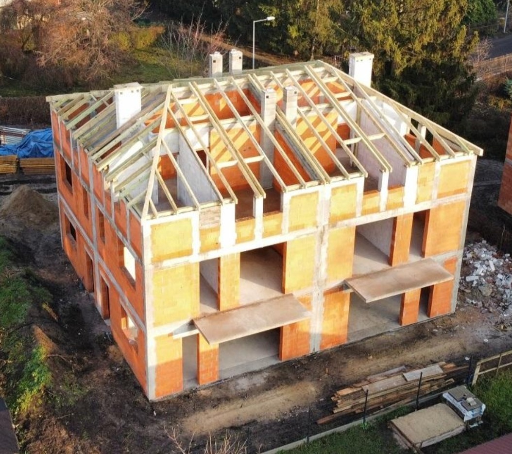 Dwukondygnacyjny dom w trakcie budowy z pustaków ceramicznych i drewnianą konstrukcją dachu, widok z góry, teren budowy z materiałami i zielenią w tle.