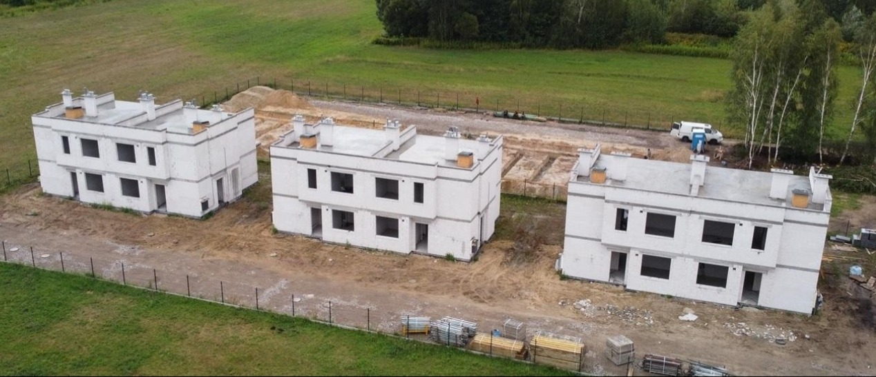 Trzy identyczne, dwukondygnacyjne domy w stanie surowym otwartym, z kominami i widocznymi fundamentami pod kolejne budynki. Ujęcie z góry.
