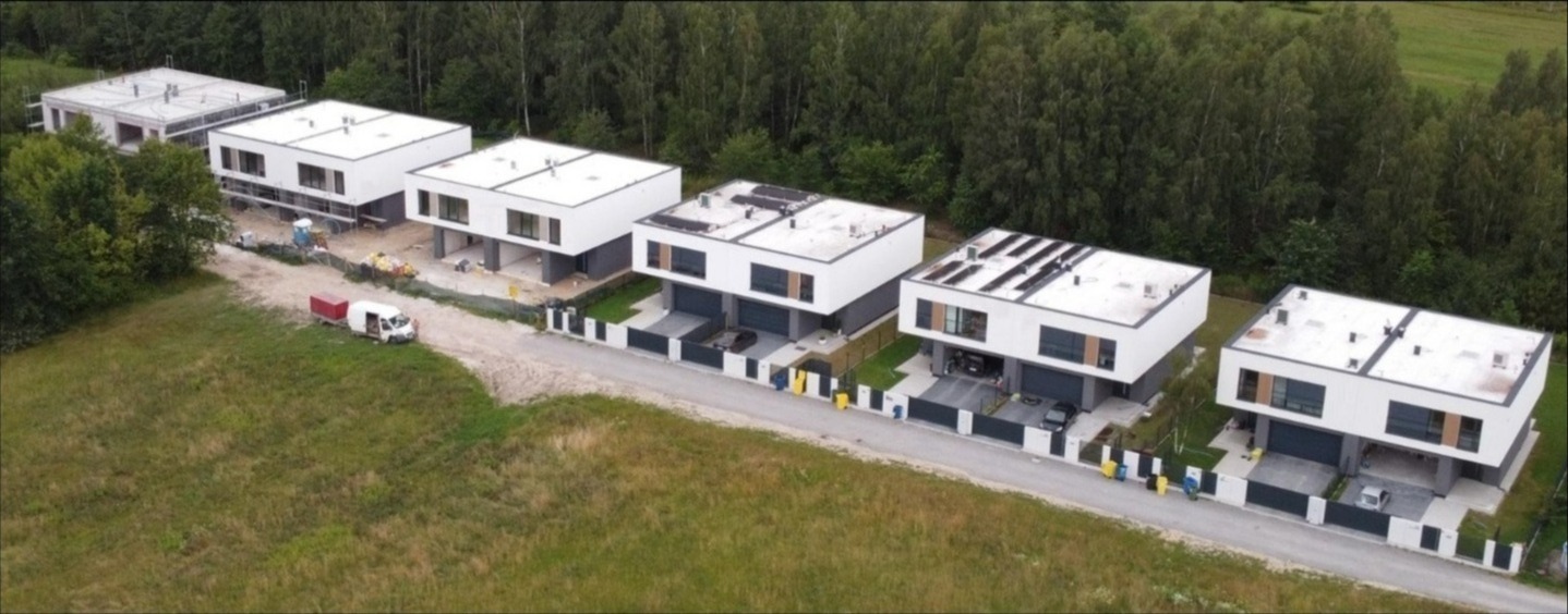 Panorama z lotu ptaka: szereg nowoczesnych, dwukondygnacyjnych domów w minimalistycznym stylu, od stanu surowego do wykończonych, na tle zielonego lasu i łąki.