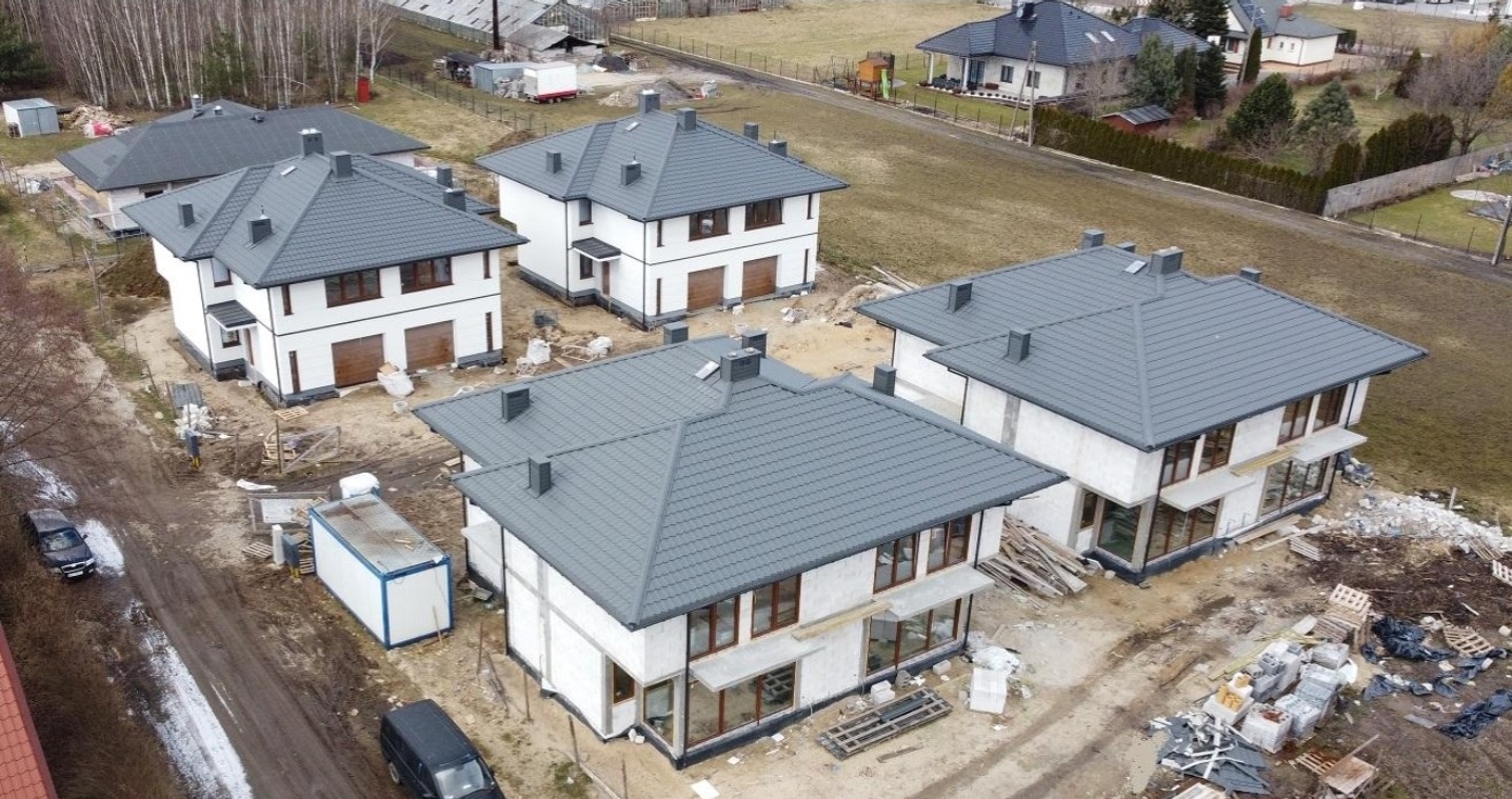 Osiedle domów w stanie surowym zamkniętym, widok z drona. Domy z szarą dachówką, białe ściany, brązowe okna. Na działkach gruz i materiały budowlane. Krajobraz podmiejski.