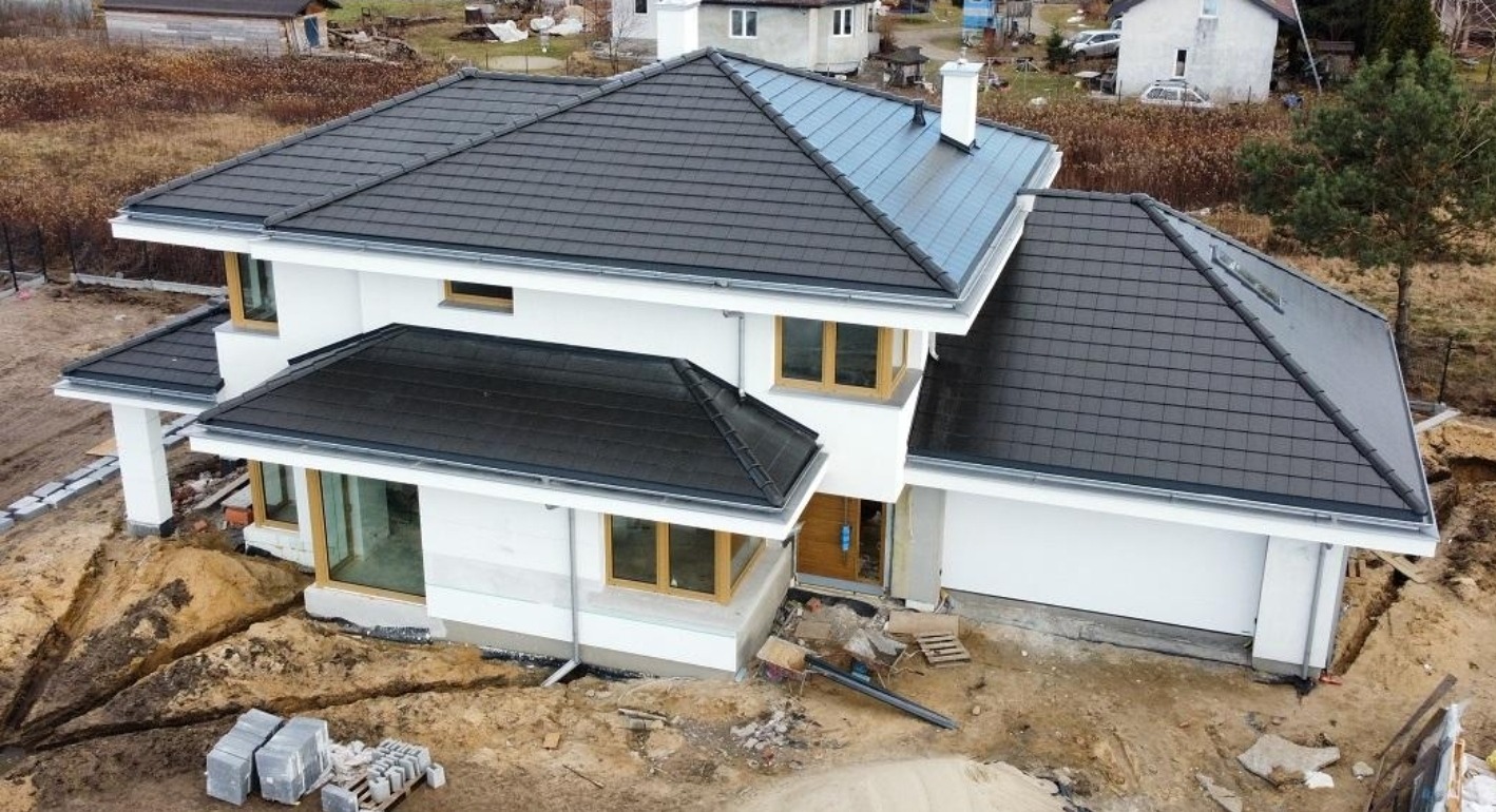 Nowoczesny, dwukondygnacyjny dom z ciemną dachówką i jasną elewacją w trakcie prac wykończeniowych. Widoczne wykopane fundamenty i materiały budowlane.