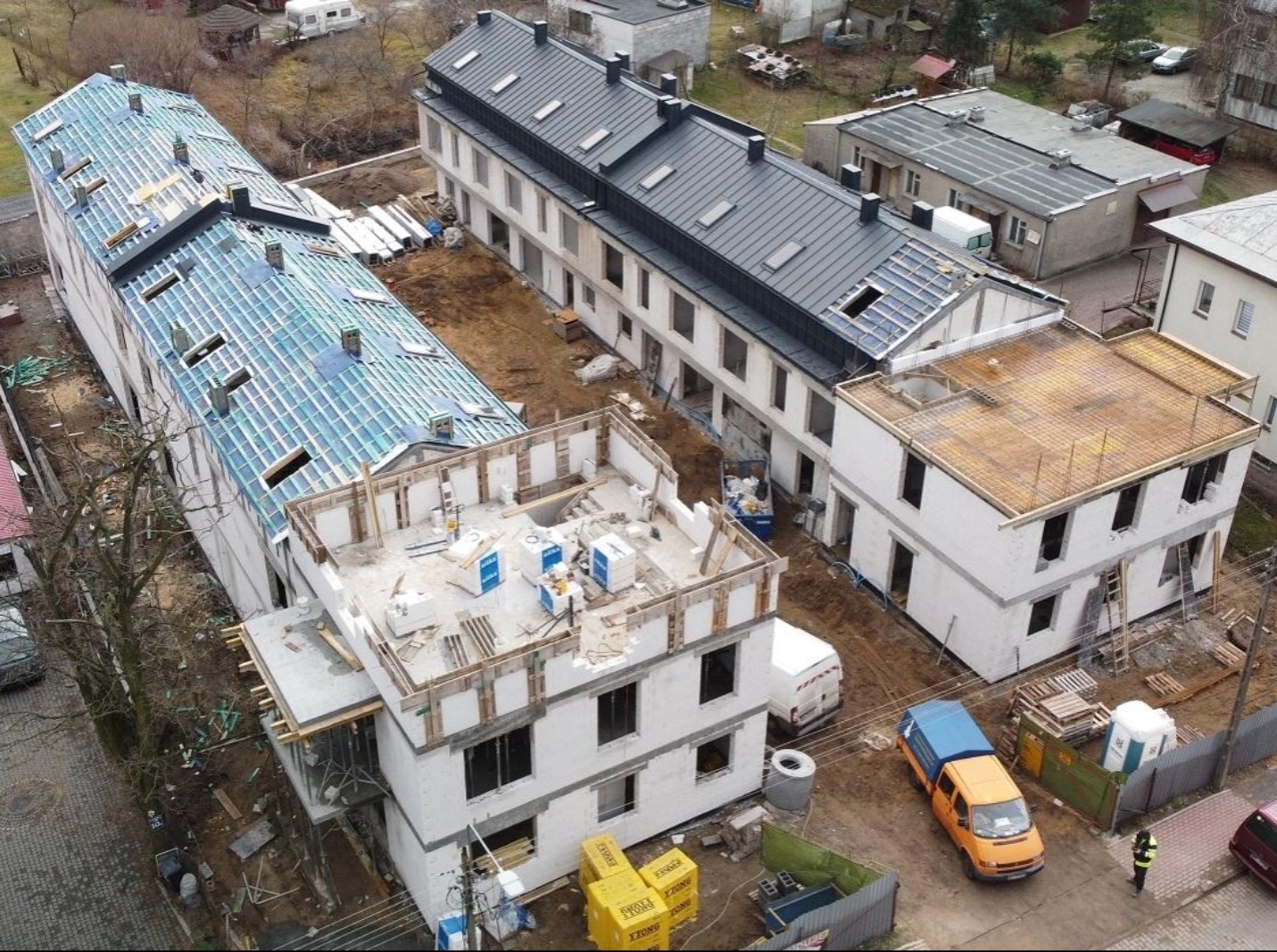 Wysoki kąt: budowa kompleksu budynków w różnym stanie ukończenia, z widocznymi elementami konstrukcyjnymi i materiałami budowlanymi, oraz pojazdami.