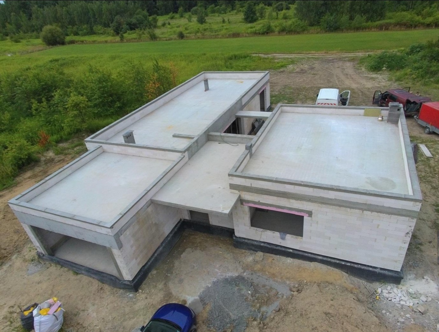 Surowy stan domu z płaskim dachem, widok z góry. Ściany z betonu komórkowego, widoczne wnęki okienne i garaż. Na dachu sprzęt budowlany i pojazdy.