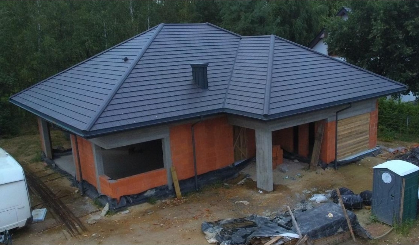 Budowa domu jednorodzinnego z ciemnym dachem i pomarańczowymi ścianami z pustaków. Częściowo widoczne deskowanie i przenośna toaleta na placu budowy.