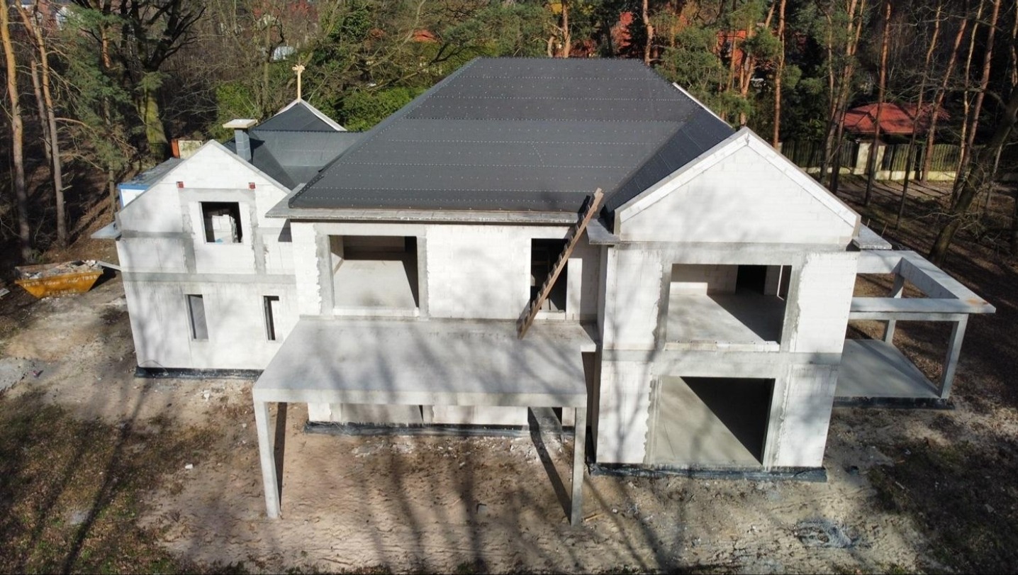 Wysoki kąt na dwukondygnacyjny, surowy dom z szarego pustaka i ciemnym dachem, otoczony drzewami. Widoczne rusztowania i otwarte przestrzenie okienne.