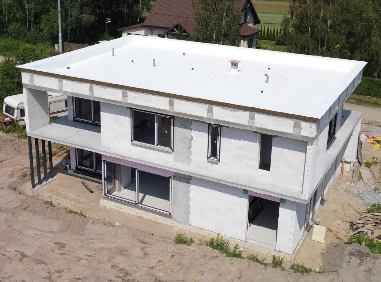 Dwukondygnacyjny budynek w stanie surowym zamkniętym z płaskim dachem i dużymi oknami, widziany z lotu ptaka na tle zieleni.