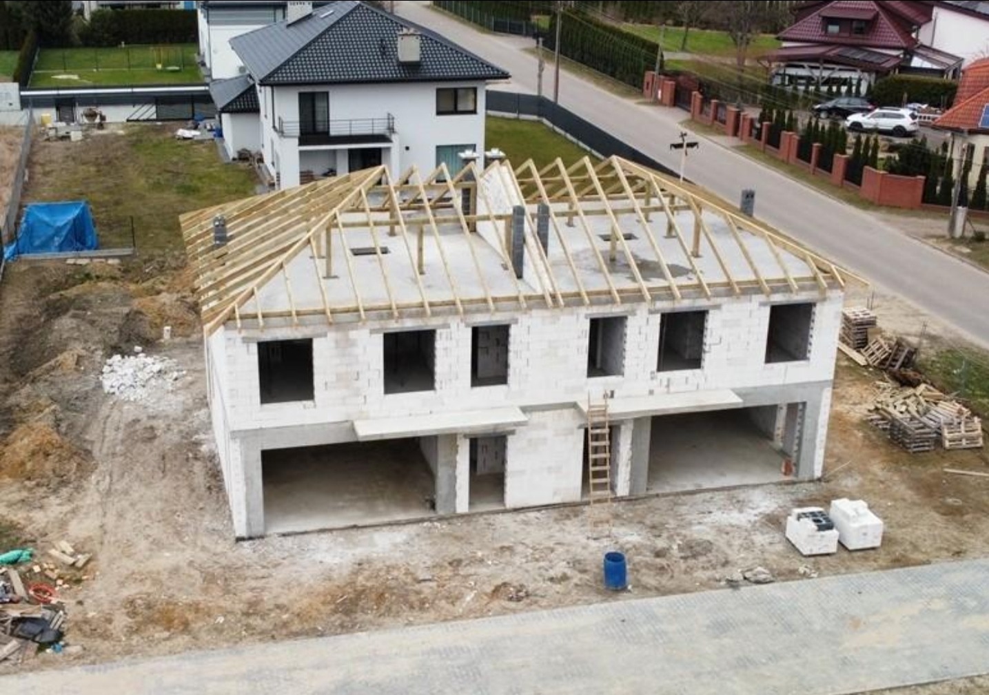 Dwukondygnacyjny dom w stanie surowym otwartym z drewnianą konstrukcją dachu, widok z góry. Na pierwszym planie gruz i materiały budowlane.