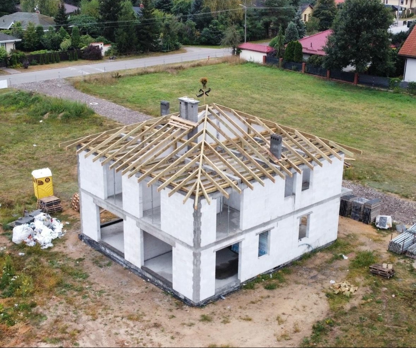 Budowa domu z białych bloczków: stan surowy otwarty z drewnianą konstrukcją dachu. Na działce widoczny przenośny WC i materiały budowlane.