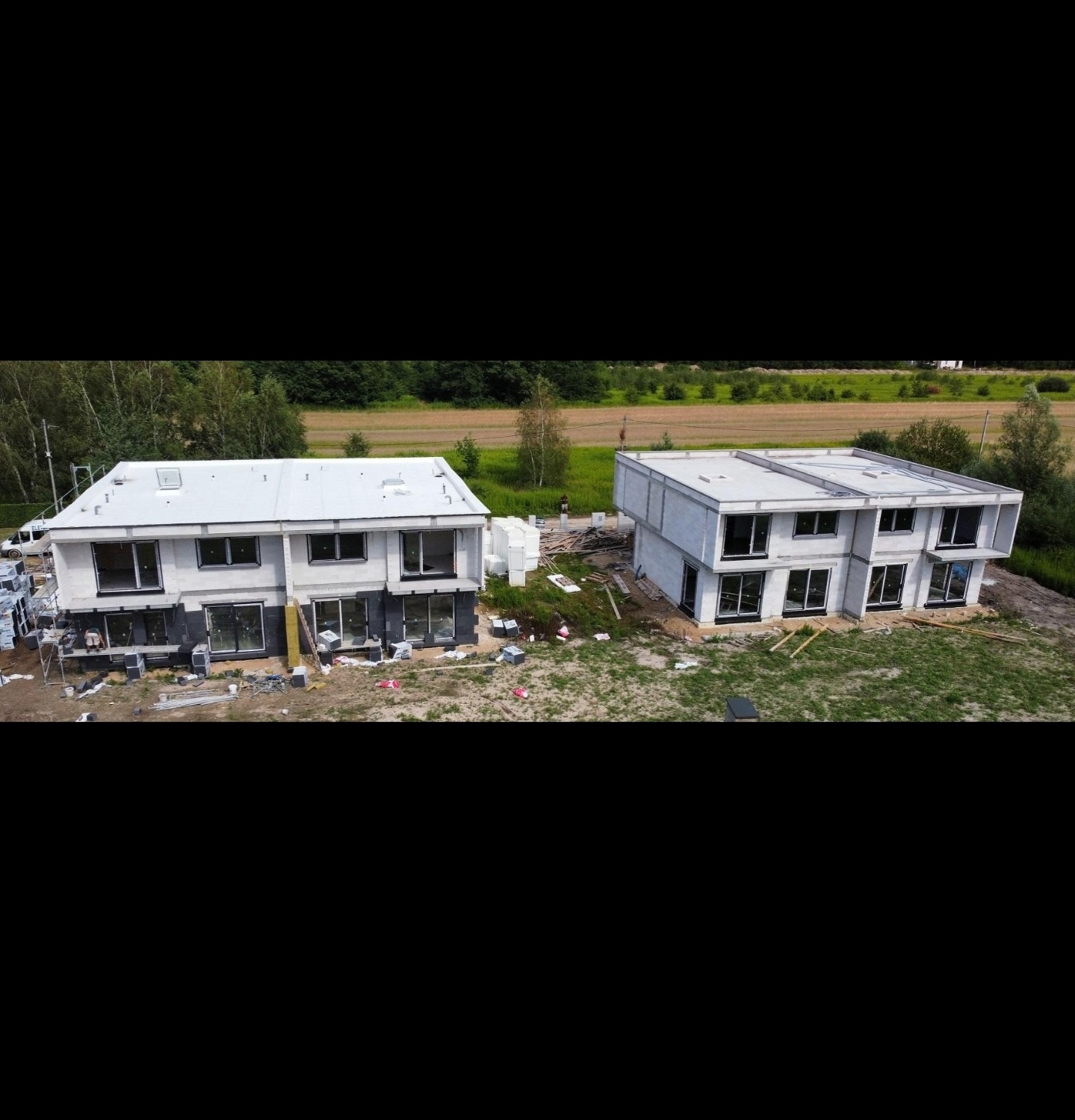 Dwa nowoczesne, dwukondygnacyjne domy w stanie surowym zamkniętym, z widocznymi elementami ocieplenia i montażu okien. Ujęcie z drona, w tle zielone pola i las.