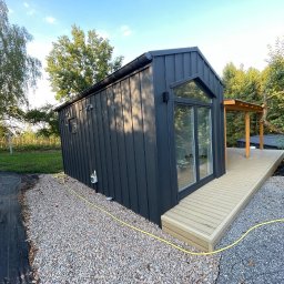 D&A Hyggedom Sp.z o.o. - Czarna, nowoczesna bryła domu modułowego z dużym oknem i drewnianym tarasem, otoczona kamiennym podłożem i zielenią. Widok z boku, w słoneczny dzień.