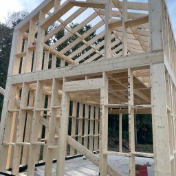 D&A Hyggedom Sp.z o.o. - Surowa konstrukcja szkieletowa domu w trakcie budowy, widok z dołu na drewniane belki i słupy, na fundamencie z zieloną izolacją. Błękitne niebo w tle.