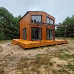 D&A Hyggedom Sp.z o.o. - Nowoczesny dom modułowy z drewnianą elewacją i tarasem w Markach, otoczony zielenią. Charakterystyczna pergola nad tarasem i duże okna.