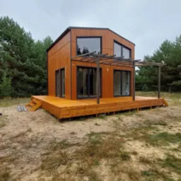 Nowoczesny dom modułowy z drewnianą elewacją i tarasem w Markach, otoczony zielenią. Charakterystyczna pergola nad tarasem i duże okna.