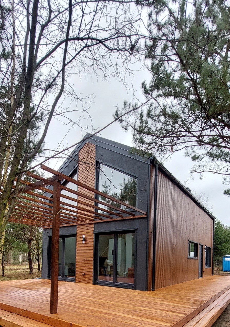 Nowoczesny dom szkieletowy z drewnianą elewacją i tarasem. Duże okna z czarnymi ramami. Pergola nad tarasem. Tło: las i pochmurne niebo.
