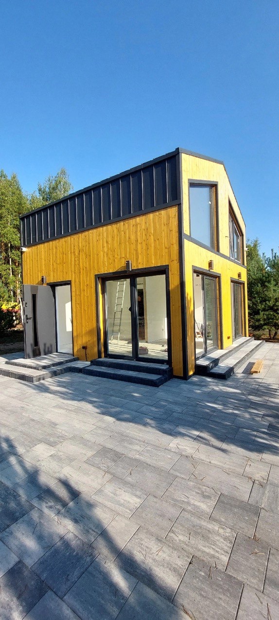 Żółty dom modułowy z czarnym dachem na tle zieleni, widok z zewnątrz. Szare, kamienne schody prowadzą do przeszklonych drzwi.
