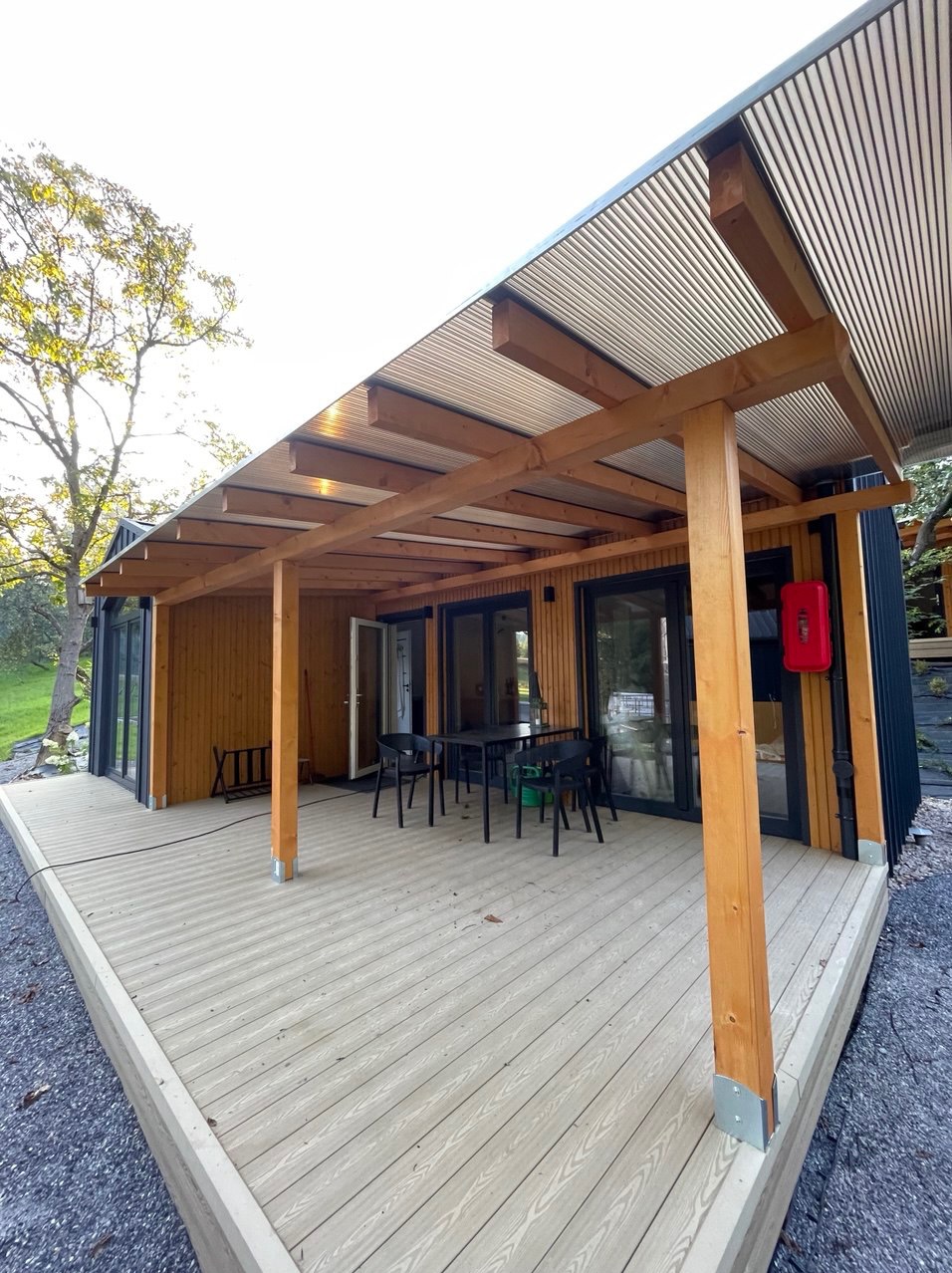 Nowoczesny dom modułowy z drewnianą elewacją i zadaszonym tarasem. Na tarasie ustawiony jest stół z krzesłami. Widoczne drewniane słupy i metalowe zadaszenie.