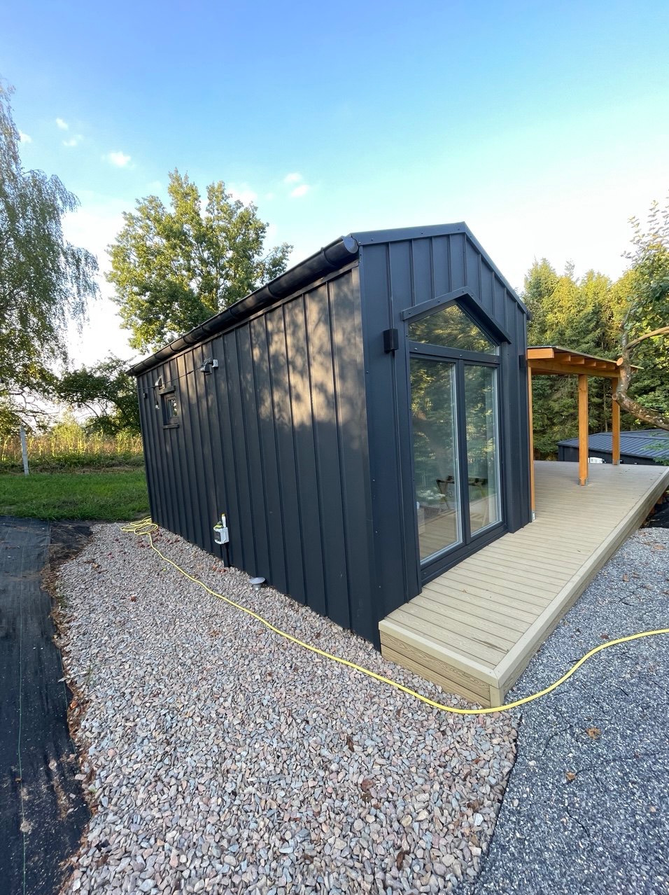 Czarna, nowoczesna bryła domu modułowego z dużym oknem i drewnianym tarasem, otoczona kamiennym podłożem i zielenią. Widok z boku, w słoneczny dzień.