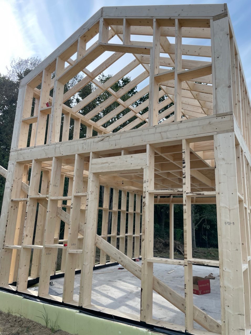 Surowa konstrukcja szkieletowa domu w trakcie budowy, widok z dołu na drewniane belki i słupy, na fundamencie z zieloną izolacją. Błękitne niebo w tle.