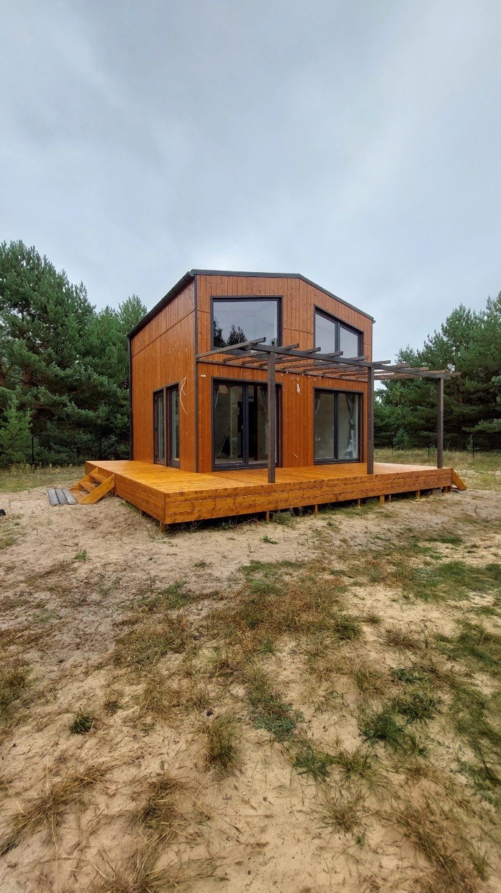 Nowoczesny dom modułowy z drewnianą elewacją i tarasem w Markach, otoczony zielenią. Charakterystyczna pergola nad tarasem i duże okna.