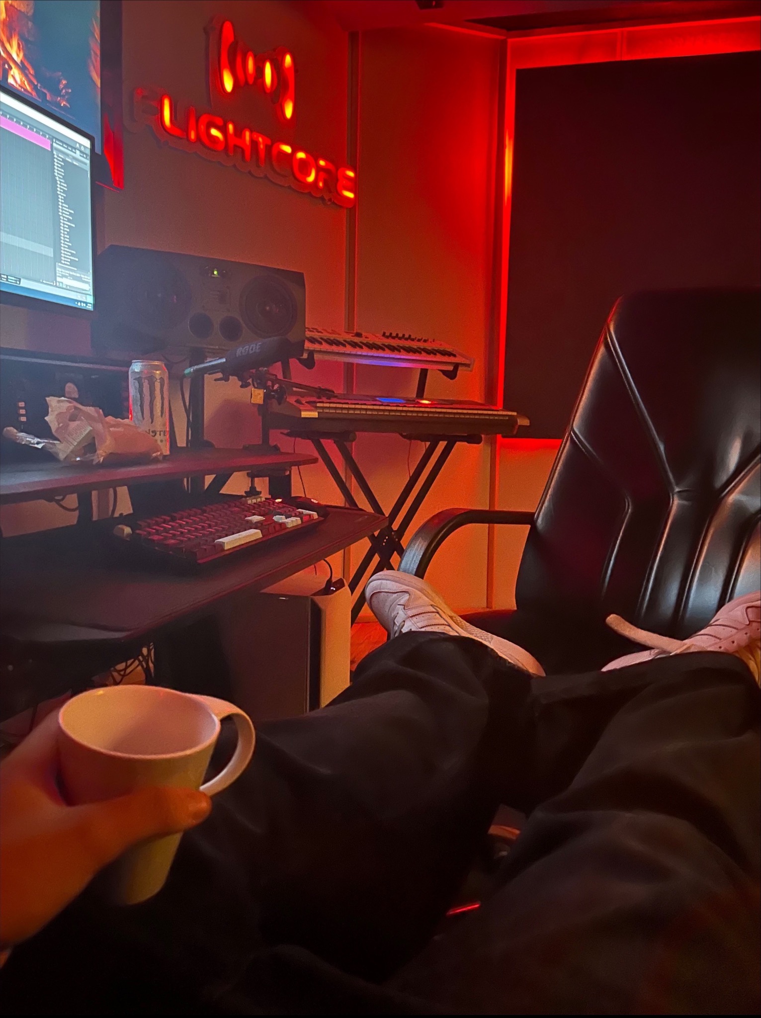 Osoba z kubkiem w fotelu w studiu nagrań z neonem 'Flightcore', klawiaturą, monitorem i mikrofonem Rode. Czerwone oświetlenie podkreśla kreatywną atmosferę.