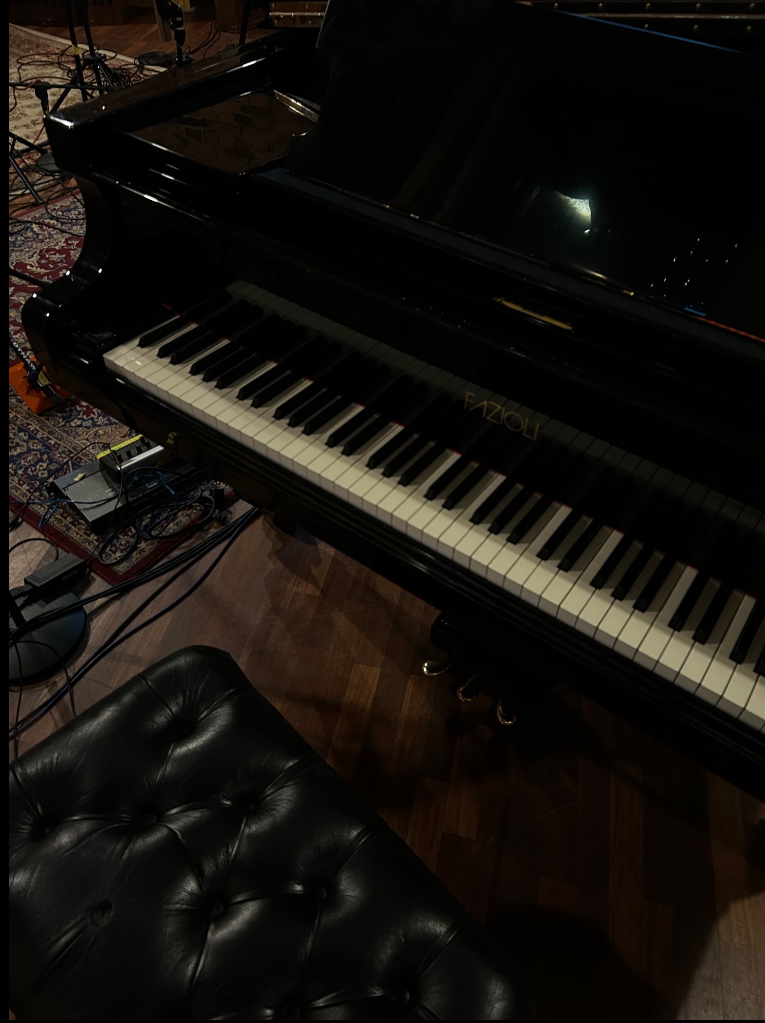 Czarny fortepian Fazioli z czarnym, skórzanym siedziskiem w studio nagrań. Widoczny dywan i sprzęt audio.