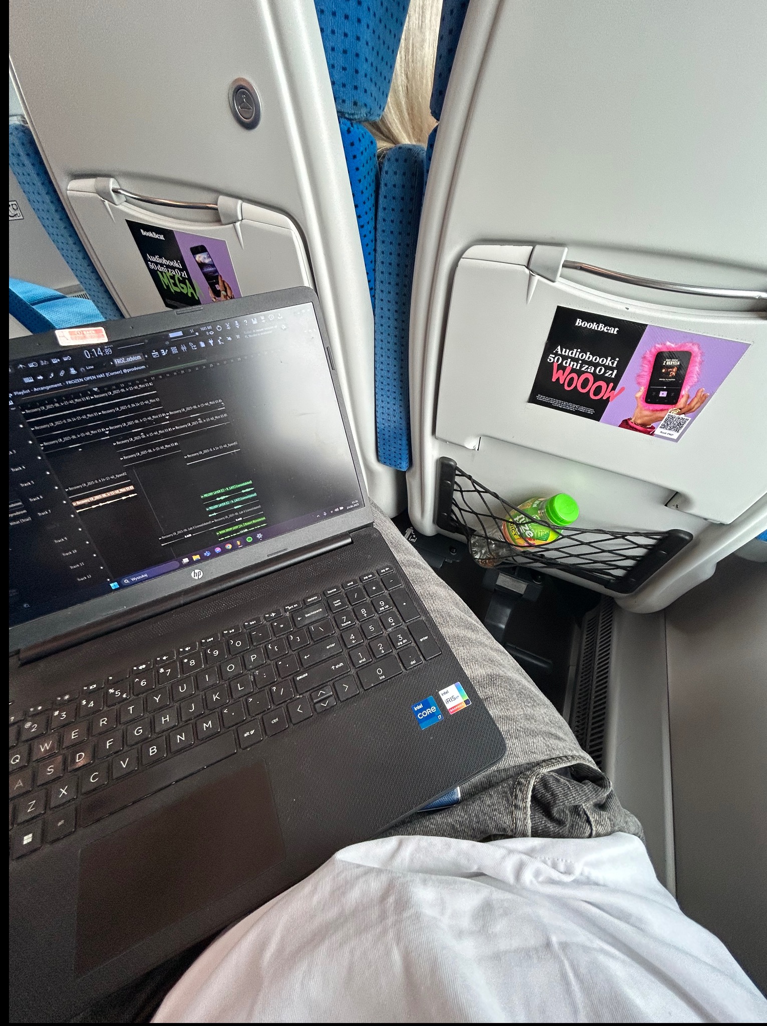 Laptop z otwartym programem DAW w pociągu, butelka wody w siatce. Widoczny ekran z interfejsem edycji audio. Praca w podróży, mobilne studio nagrań?