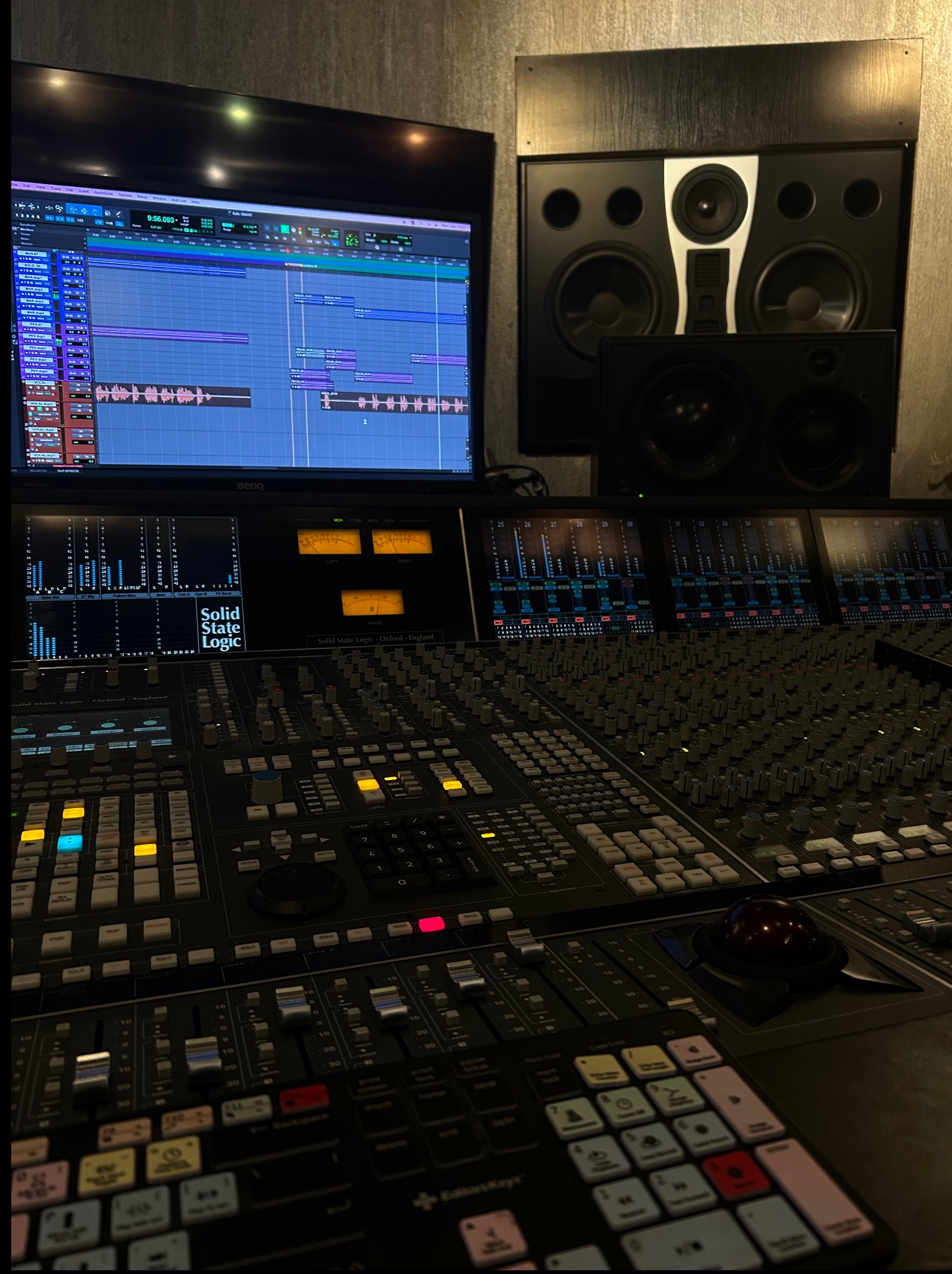 Profesjonalny stół mikserski Solid State Logic z monitorem DAW w studio nagrań. Widoczne głośniki studyjne. Oświetlenie ambientowe.