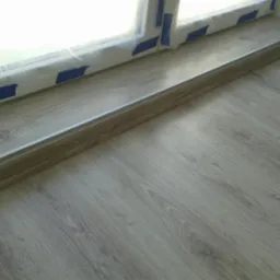 Ułożone panele podłogowe w odcieniach szarości z widocznym oknem zabezpieczonym folią i niebieską taśmą malarską.