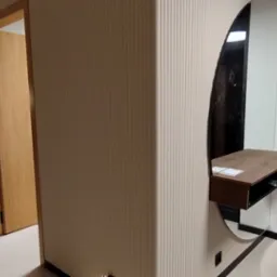 Nowoczesny przedpokój z owalnym lustrem i konsolą. Ściana z beżowymi pionowymi żłobieniami, drzwi z jasnego drewna, podłoga w odcieniach beżu. Minimalistyczny design.