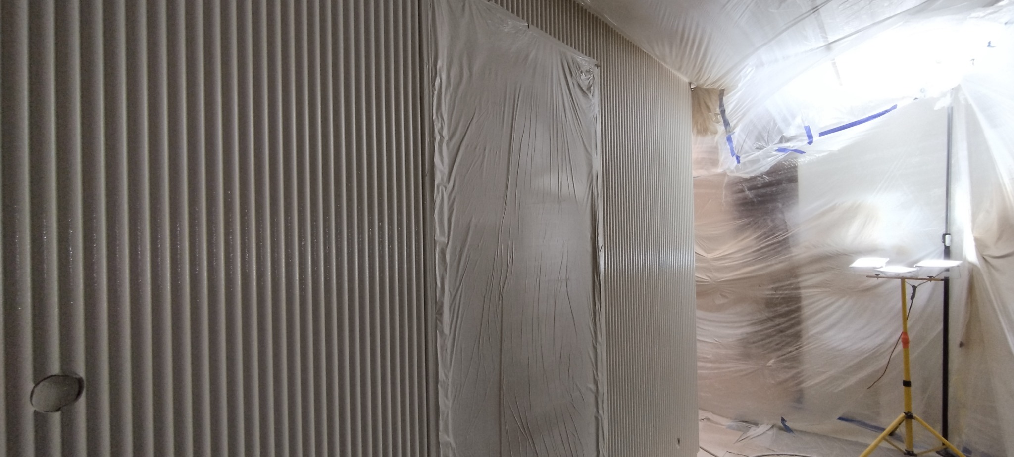 Ściana z dekoracyjnymi pionowymi żłobieniami, częściowo zakryta folią ochronną, przygotowana do malowania. Oświetlenie halogenowe na statywie.