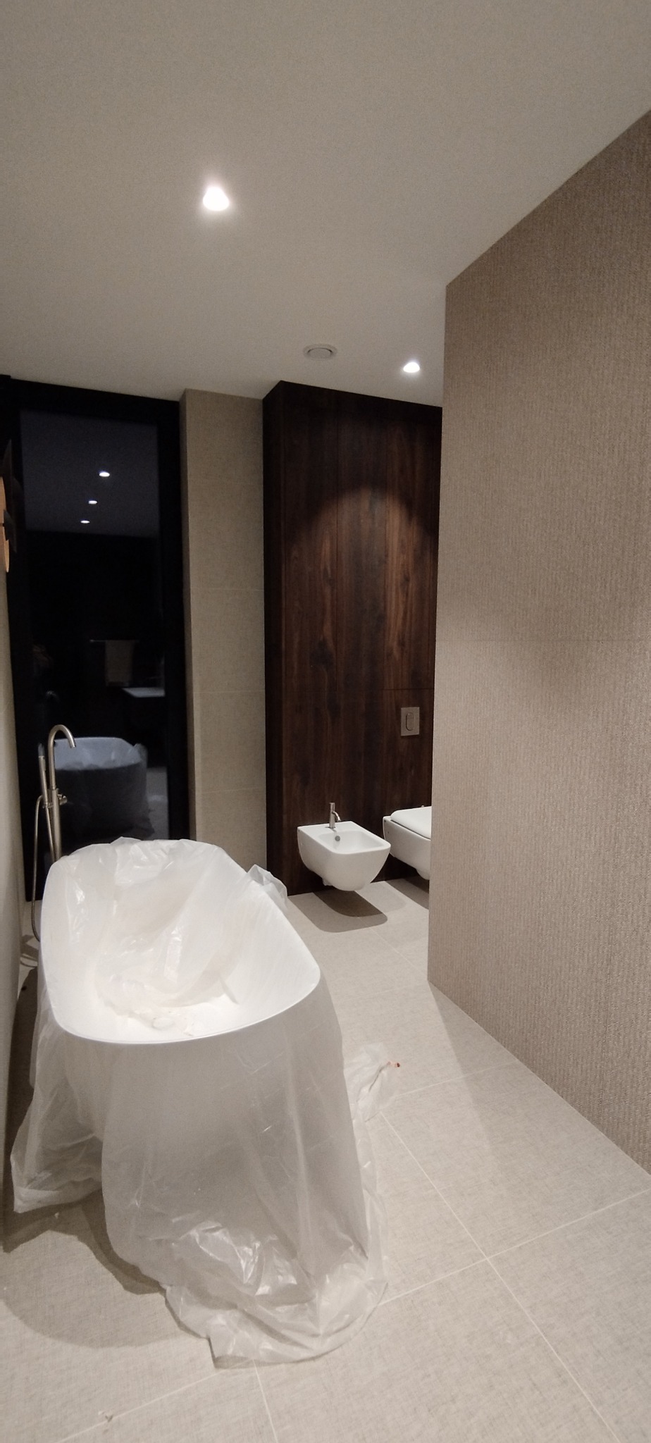 Nowoczesna łazienka w trakcie wykończenia: wolnostojąca wanna zabezpieczona folią, bidet i toaleta, drewniana zabudowa. Stonowana kolorystyka, jasne płytki podłogowe.