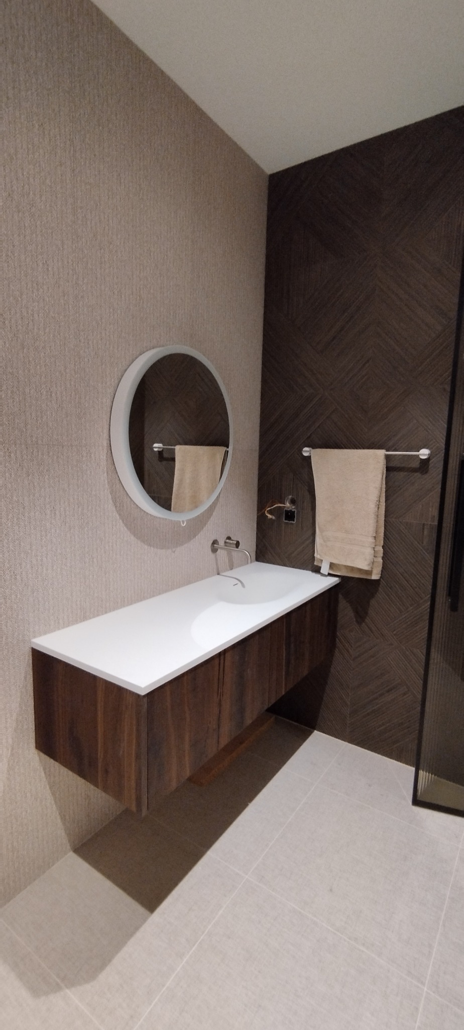 Nowoczesna łazienka z drewnianą szafką pod umywalką i okrągłym lustrem. Na ścianie geometryczne brązowe płytki i beżowa ściana z teksturą.