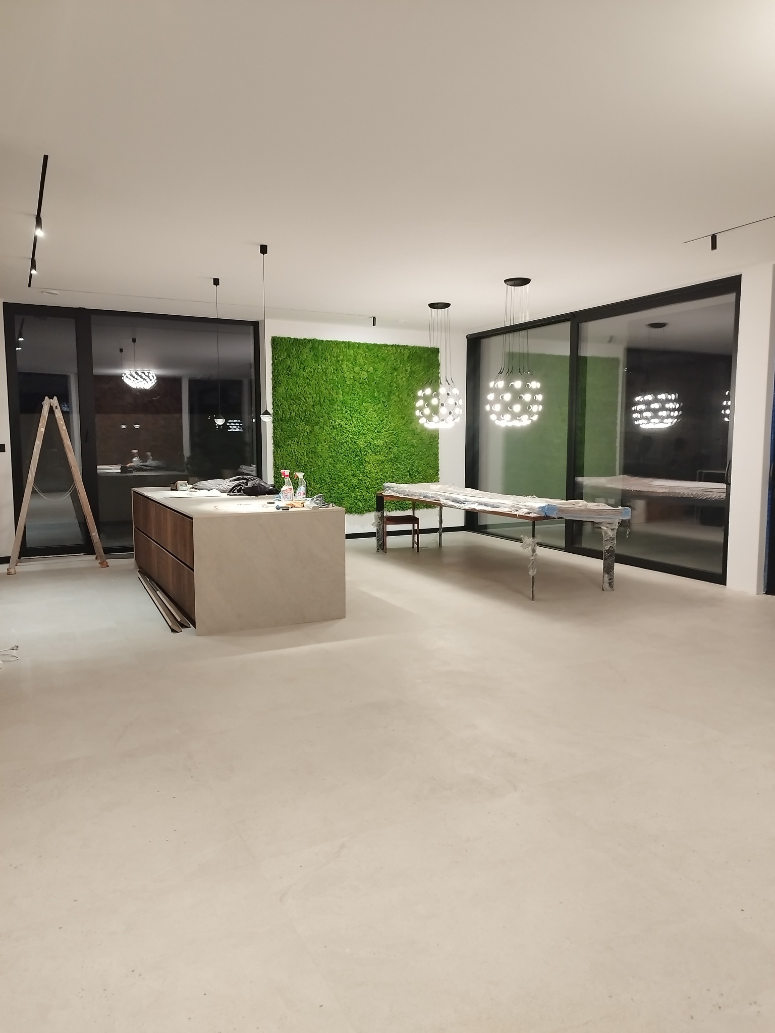 Wnętrze w trakcie wykańczania: wyspa kuchenna z drewnianymi szufladami, zabezpieczone meble, drabina i ściana z mchu. Nowoczesne lampy wiszące.