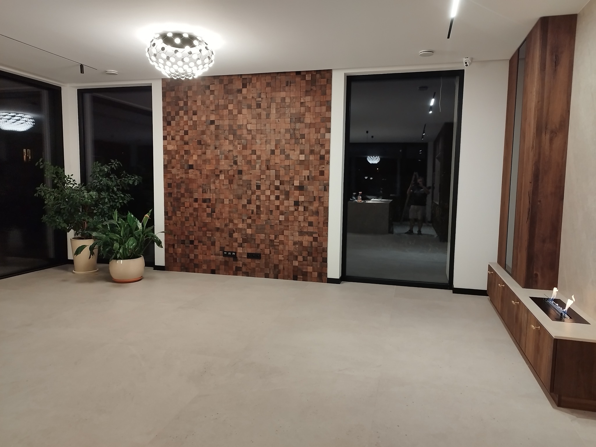 Nowoczesne wnętrze z drewnianą ścianą z kwadratów, minimalistycznym oświetleniem i roślinami doniczkowymi. Widoczne duże okna i bio-kominek.