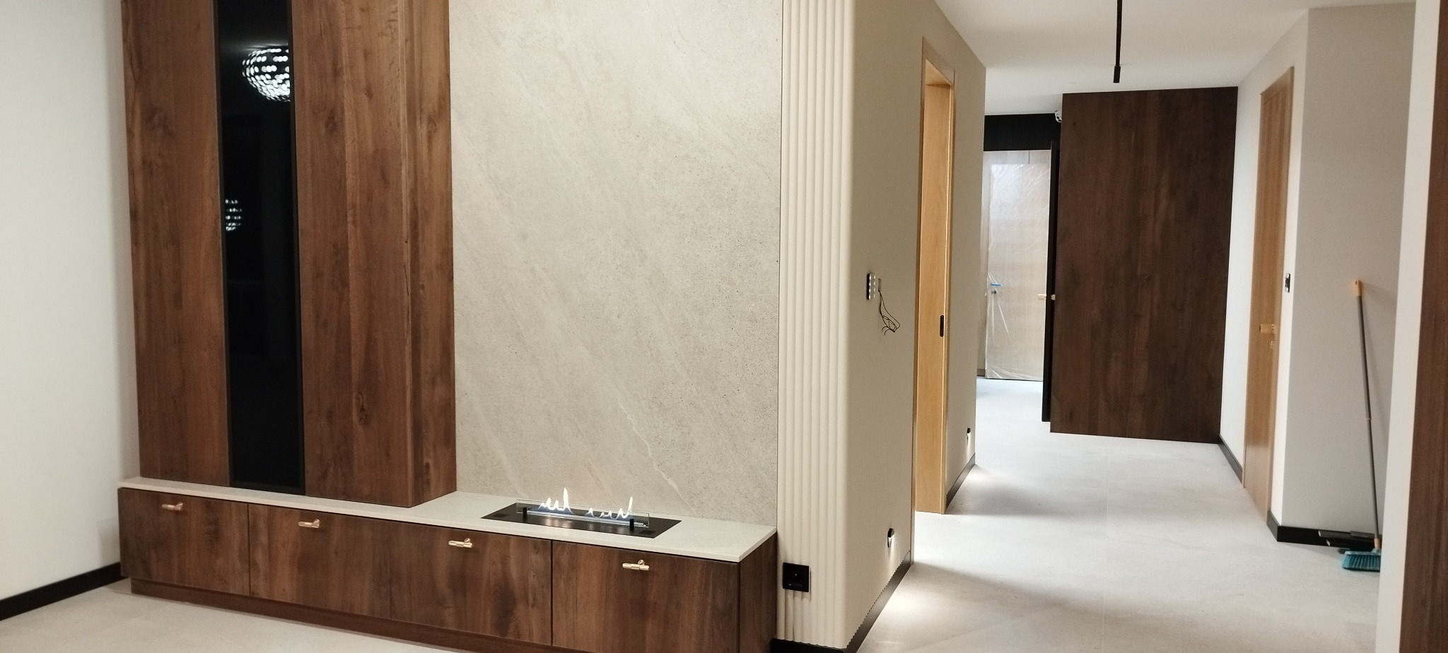 Nowoczesny salon z drewnianą zabudową, biokominkiem i beżową ścianą. Widoczne drzwi i fragment podłogi. Minimalistyczny design z dbałością o detale.