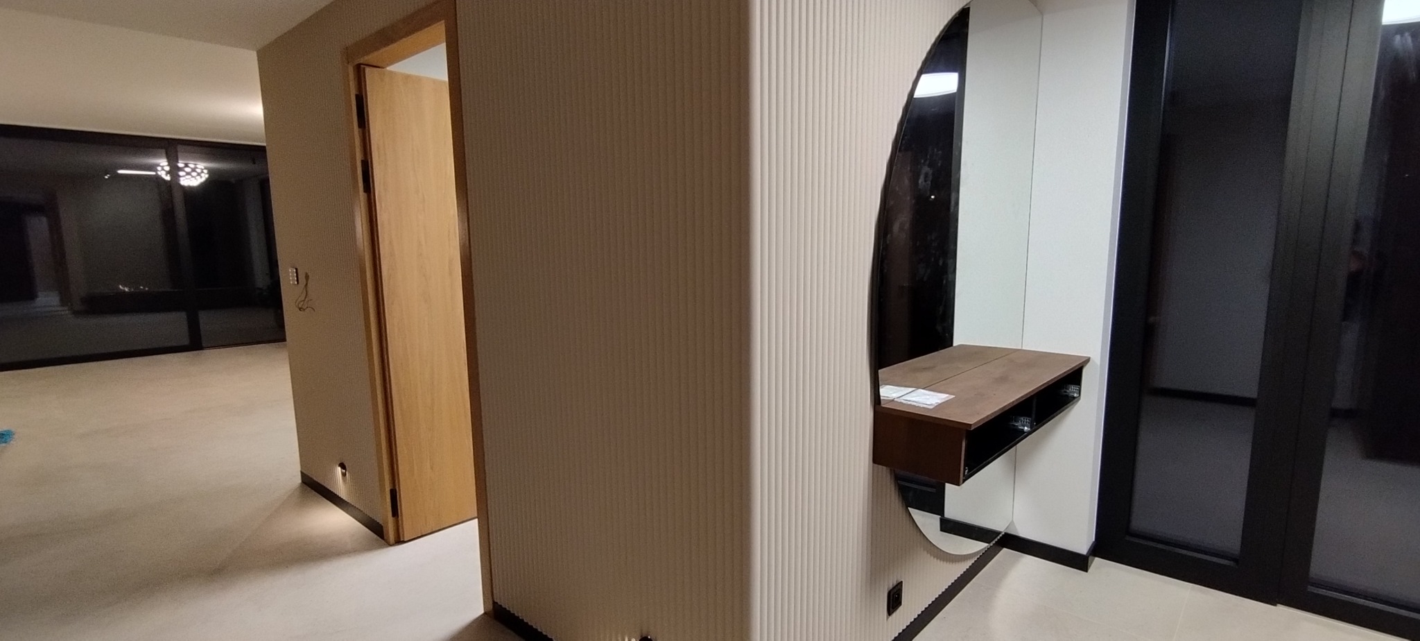 Nowoczesny przedpokój z owalnym lustrem i konsolą. Ściana z beżowymi pionowymi żłobieniami, drzwi z jasnego drewna, podłoga w odcieniach beżu. Minimalistyczny design.