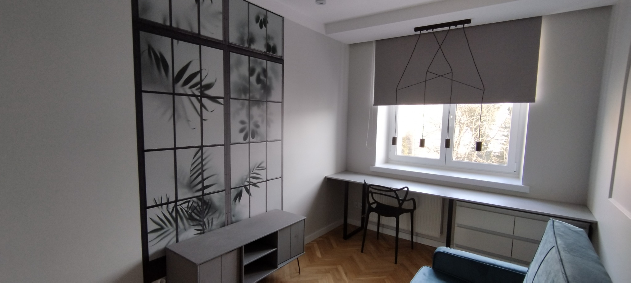 Pokój po remoncie: szara komoda, ściana z motywem roślinnym, biurko pod oknem z roletą i lampą wiszącą. Parkiet na podłodze, fragment sofy w rogu.