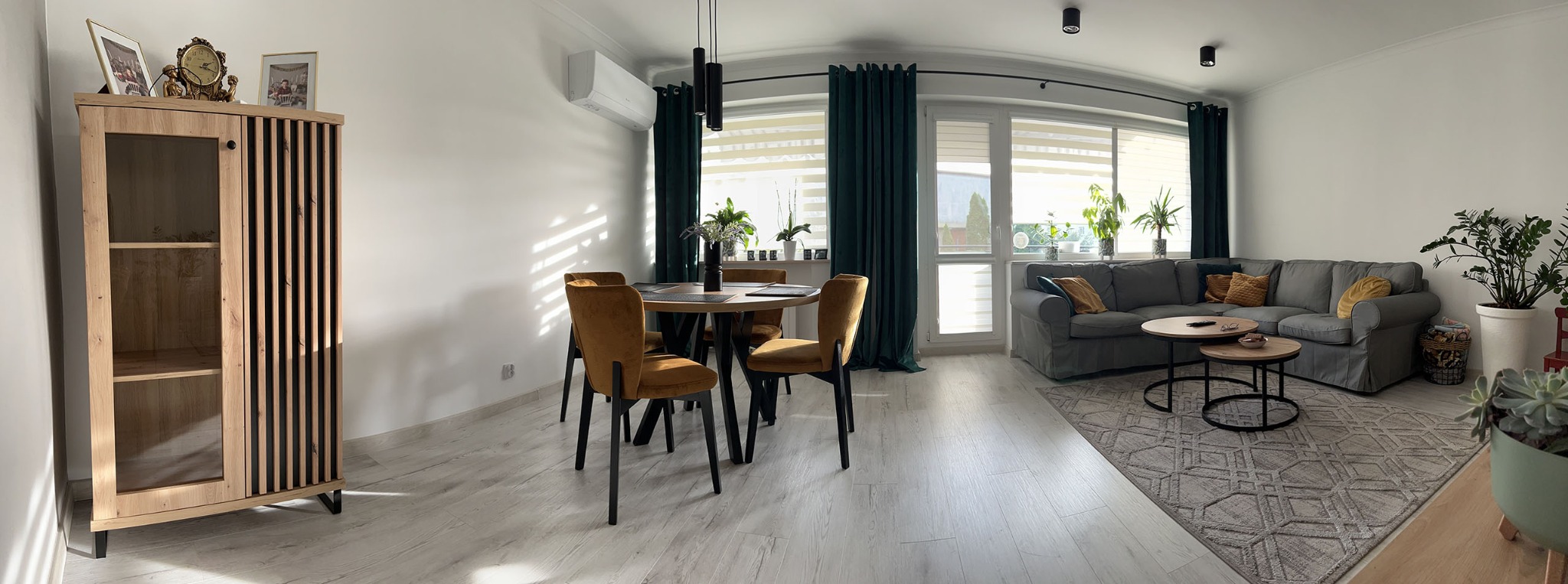 Panoramiczny widok na jasny salon z drewnianą podłogą, okrągłym stołem z musztardowymi krzesłami, szarą sofą i szafką z elementami drewna. Zielone zasłony i rośliny dodają koloru.