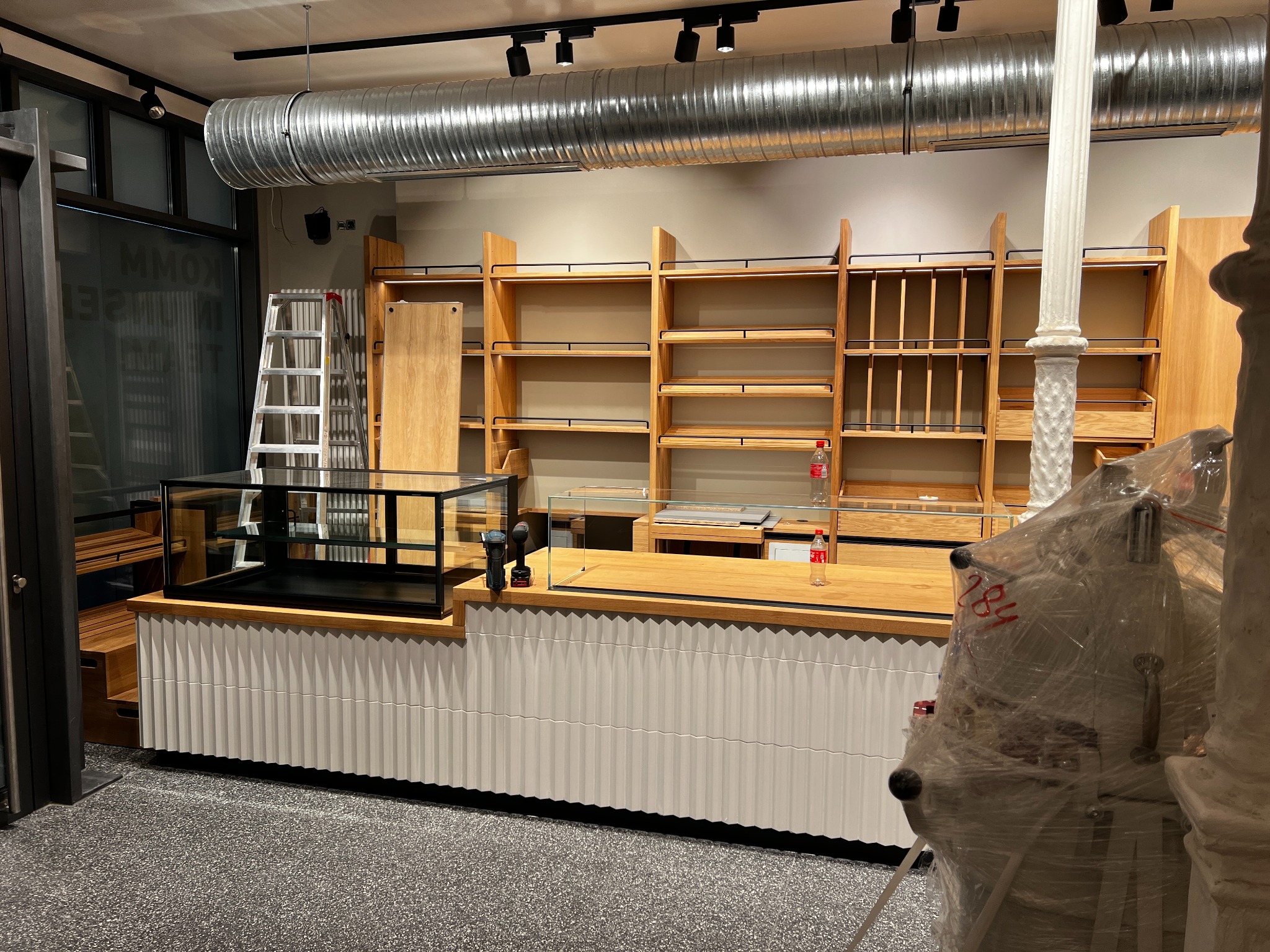 Nowe, drewniane regały sklepowe z czarnymi metalowymi elementami, biała lada z ryflowanym frontem i szklana witryna w surowym wnętrzu sklepu w trakcie wykańczania.