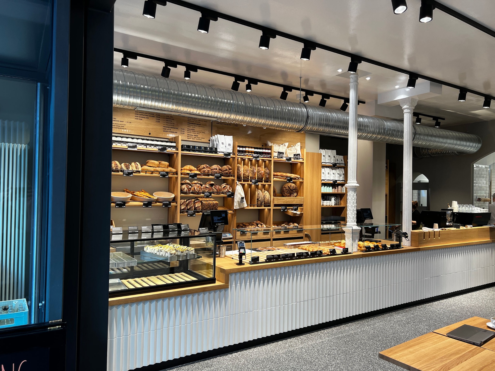 Nowoczesne wnętrze piekarni z drewnianymi półkami pełnymi pieczywa, przeszkloną ladą i białymi, żłobionymi panelami. Widoczne oświetlenie punktowe i system wentylacji.